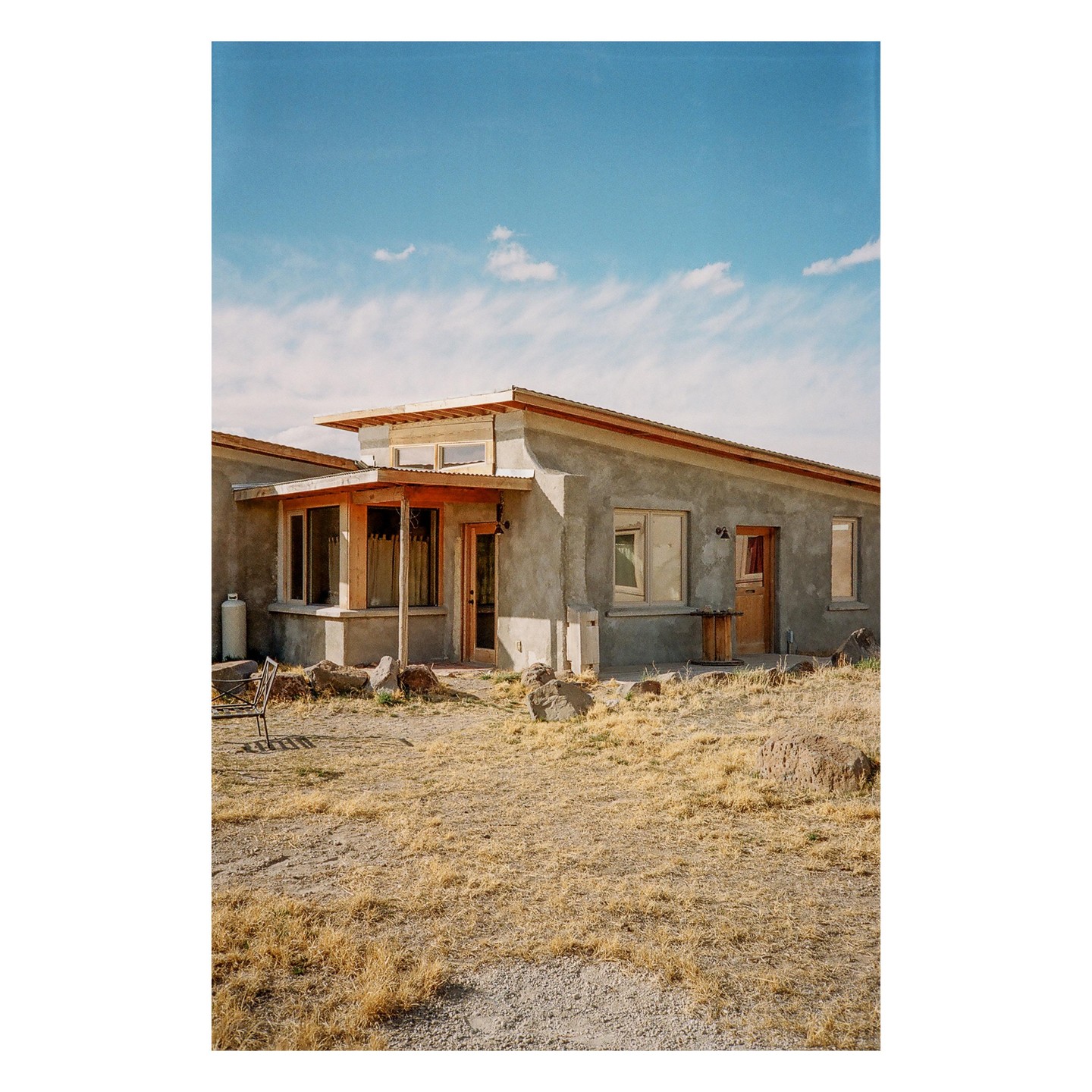 A stucco house with a wooden porch and a metal roof stands in a dry, grassy field under a blue sky with scattered clouds.