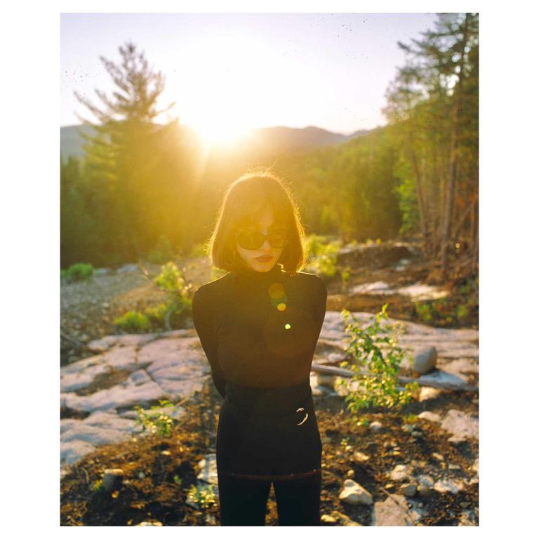 A woman with short hair and sunglasses stands in a sunlit, rocky landscape with trees and mountains in the background.