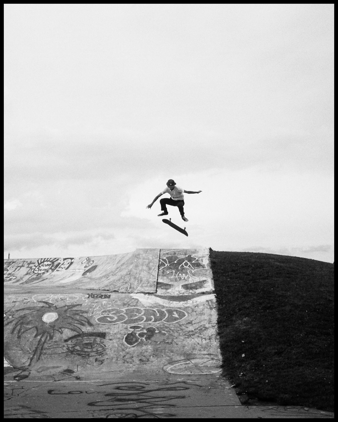 A skateboarder is captured mid-air above a graffiti-covered skate park ramp next to a grassy hill.