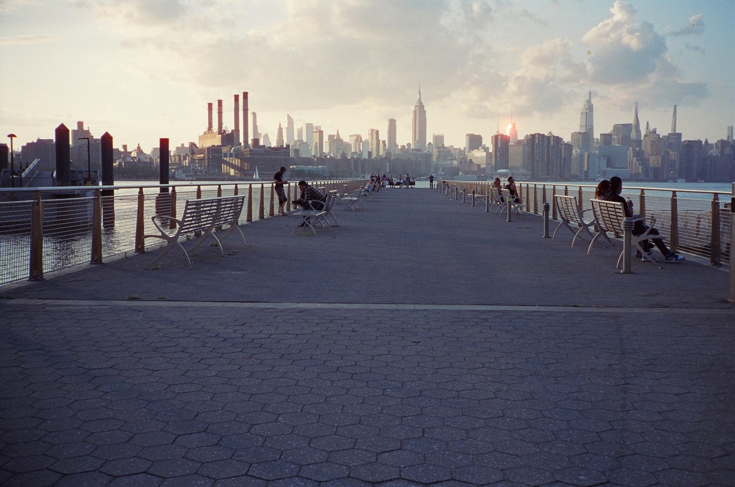 People sit on benches along a pier overlooking the new york city skyline at sunset.