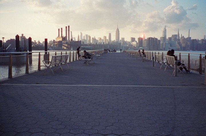 People sit on benches along a pier overlooking the new york city skyline at sunset.