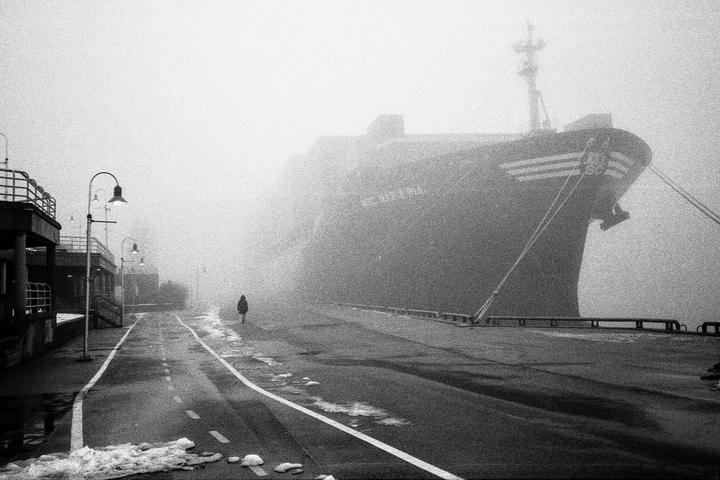 A person walks along a foggy street next to a large ship docked at a pier.