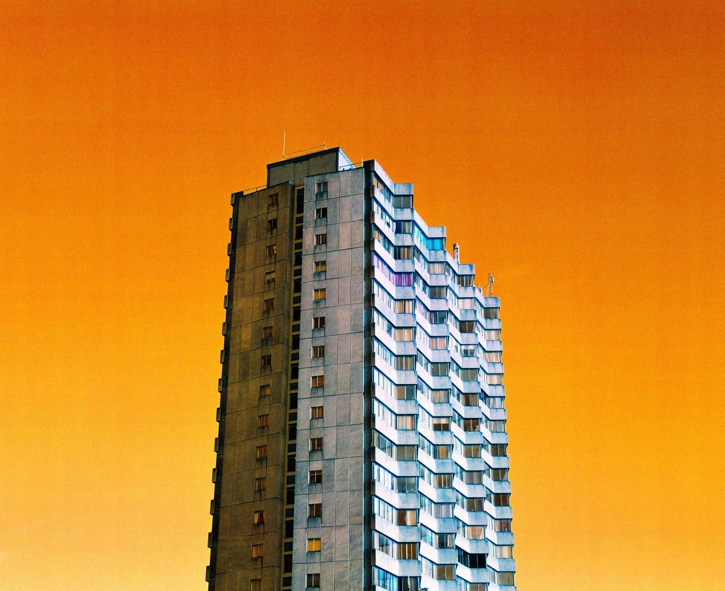 A tall, brutalist building with many windows stands against a bright orange sky.