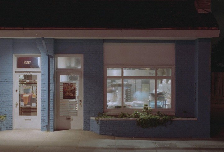 A blue brick building with two white doors and a large window displaying a bakery interior at night.
