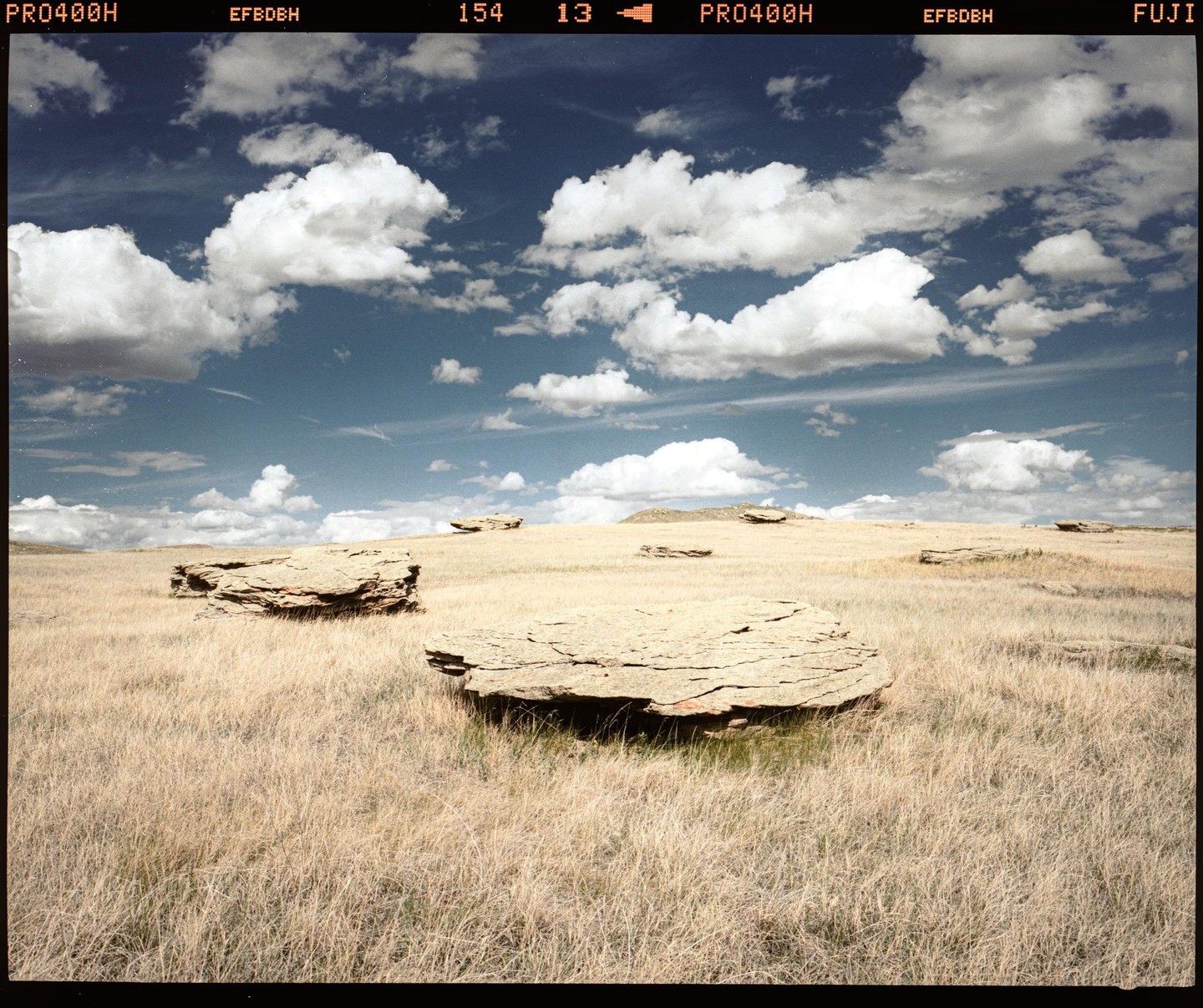 A wide-angle landscape shows a field of dry grass with several flat, round boulders under a blue sky with white clouds.