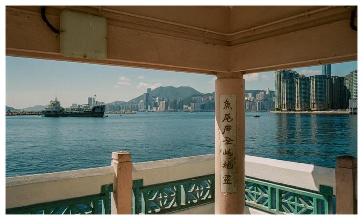 A boat is visible on the water with a city skyline and mountains in the background, framed by a structure with a railing and a pillar with Chinese characters.