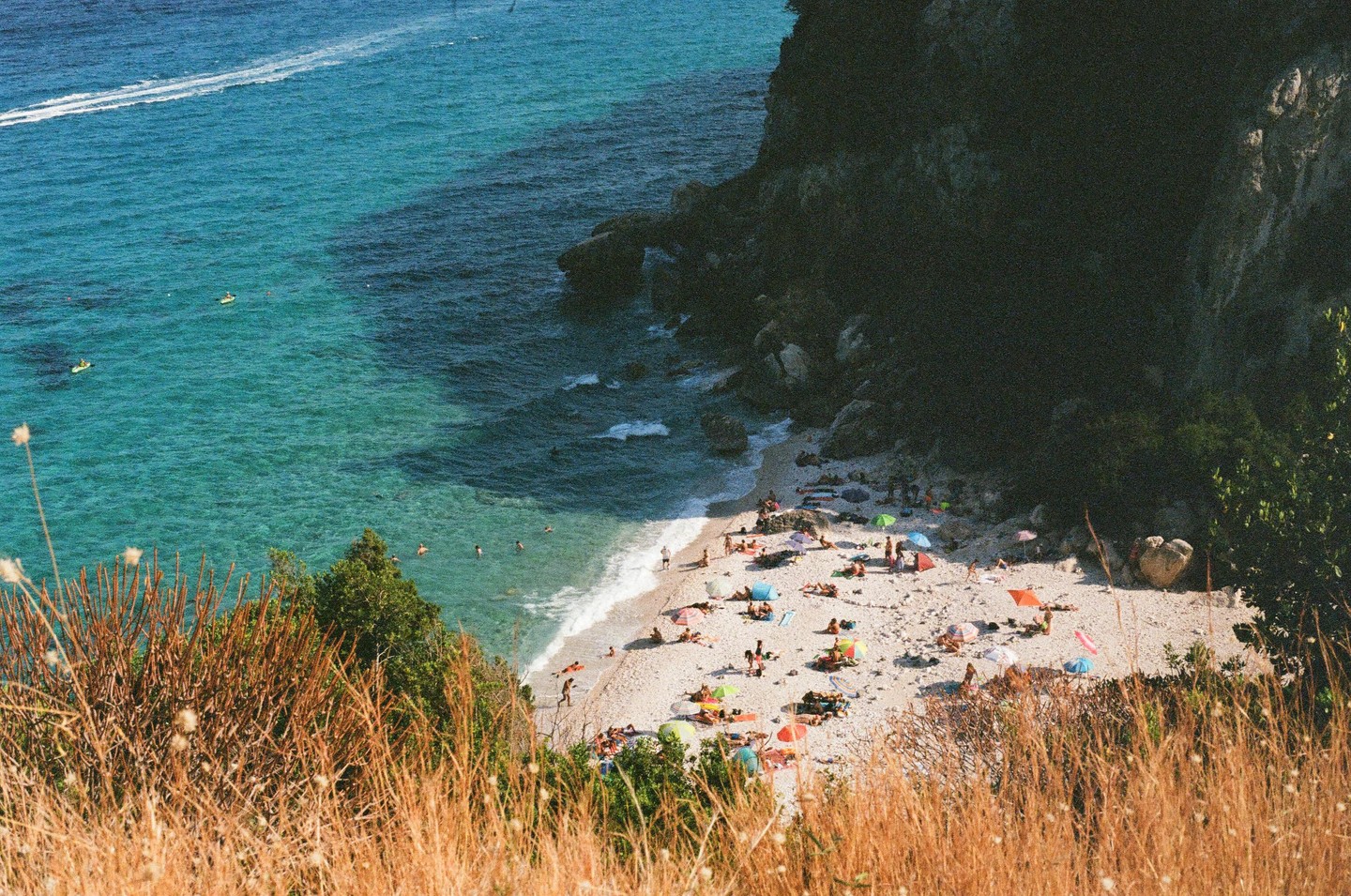 A high-angle view shows a crowded beach with many people sunbathing and swimming in the turquoise water next to a dark cliff.