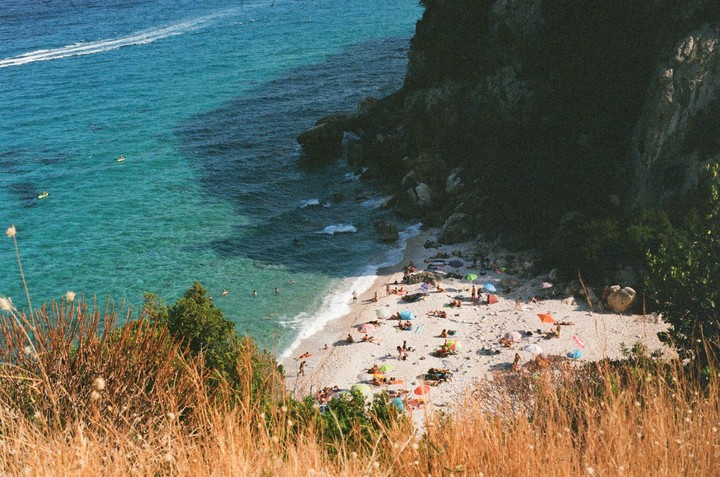 A high-angle view shows a crowded beach with many people sunbathing and swimming in the turquoise water next to a dark cliff.