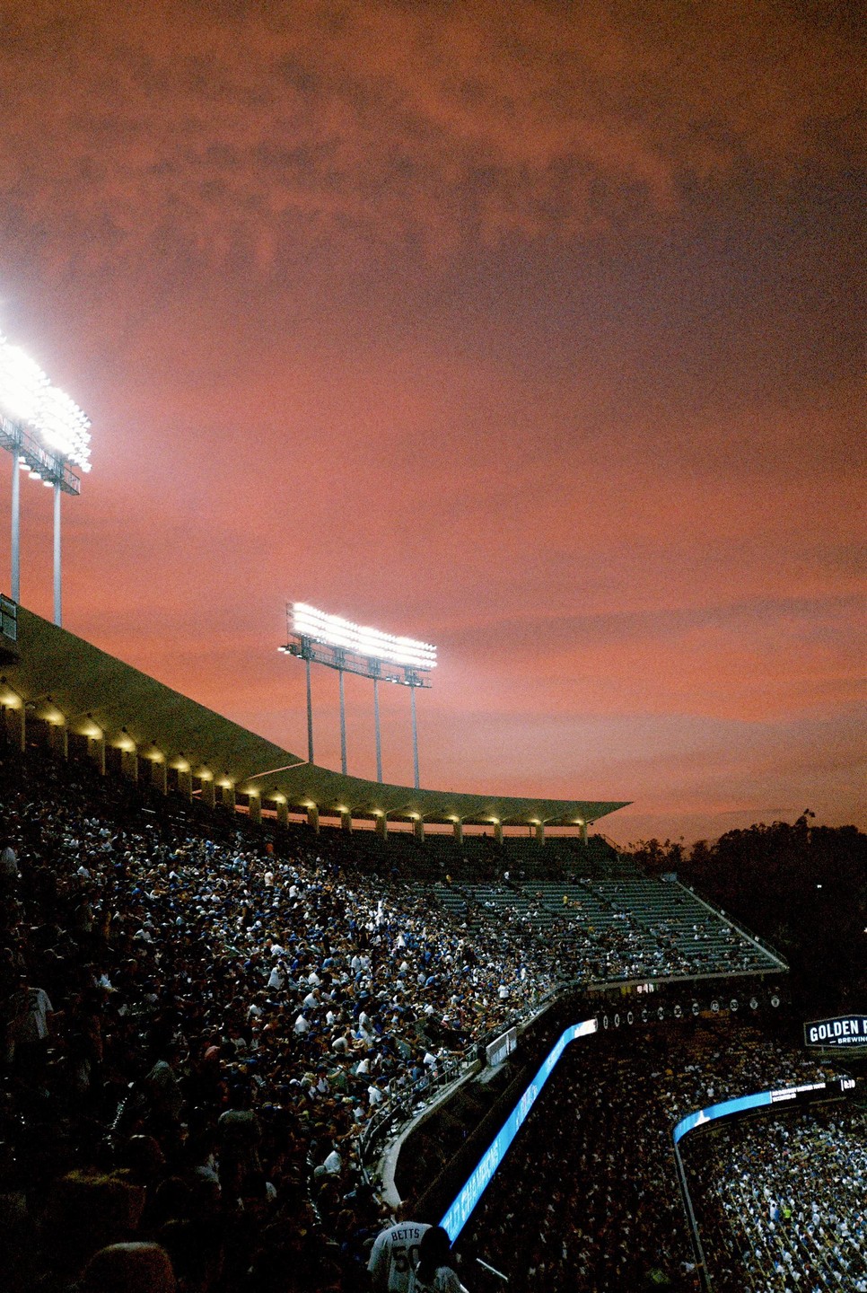 A baseball stadium is filled with spectators under a dramatic orange sky at sunset.