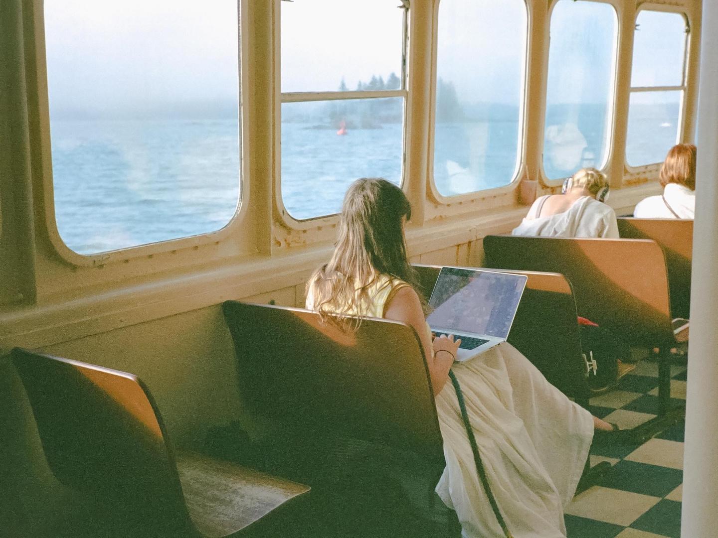 A woman sits on a ferry and uses a laptop while looking out at the water.