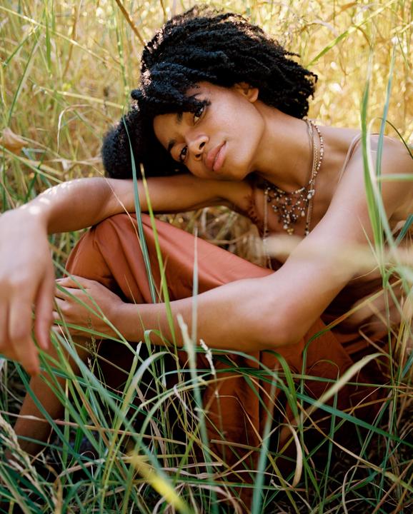 A woman with curly hair and necklaces is sitting in tall grass.