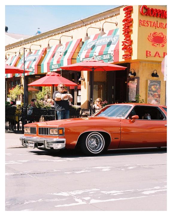 A man holds a small dog in front of a restaurant with a classic orange car parked on the street.