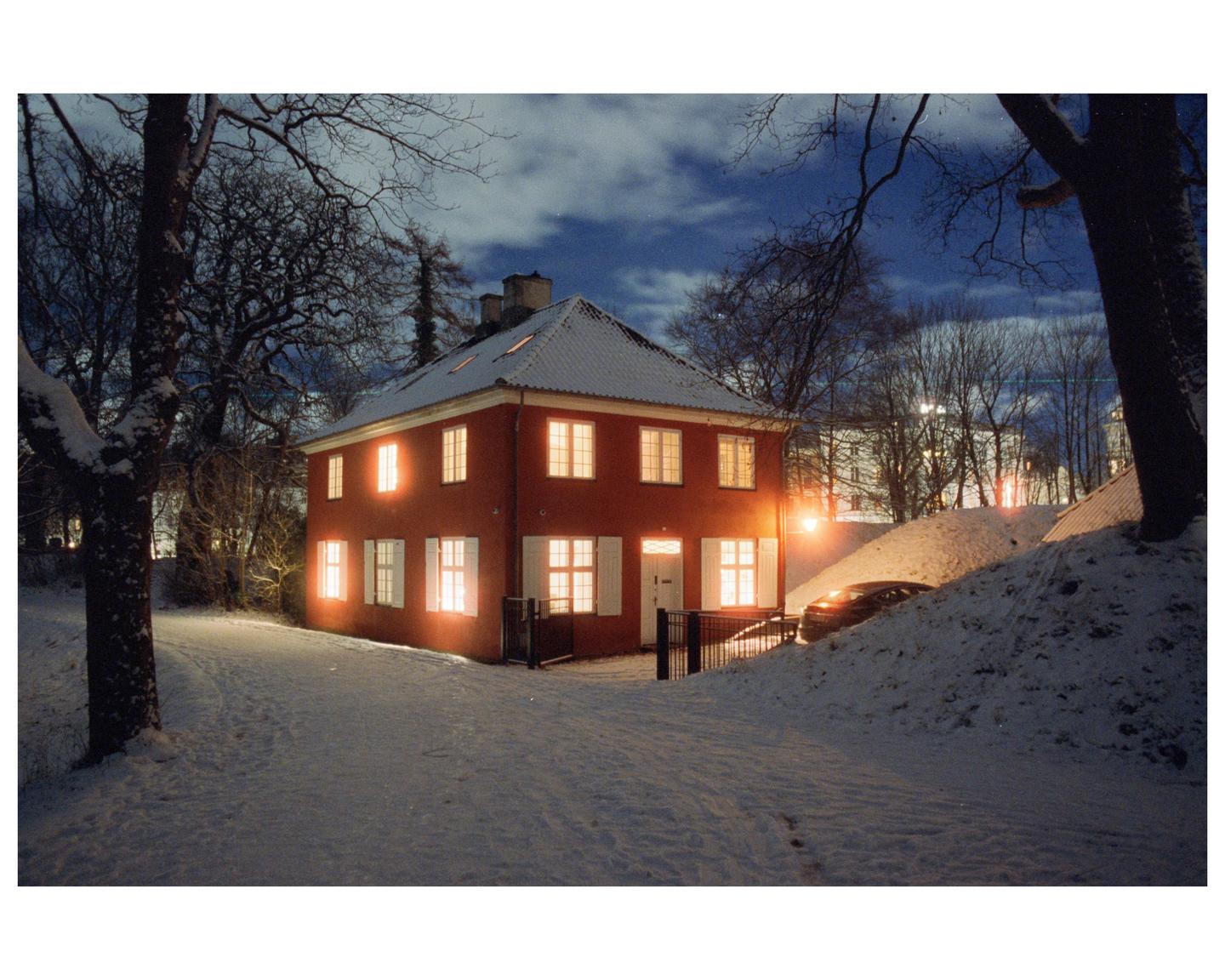 A red house with illuminated windows stands in a snowy landscape at night.