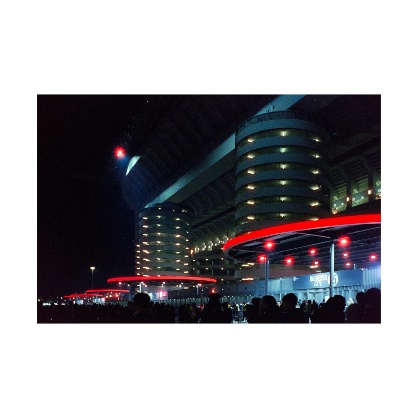 A crowd of people gathers outside a large stadium with red and white lights at night.