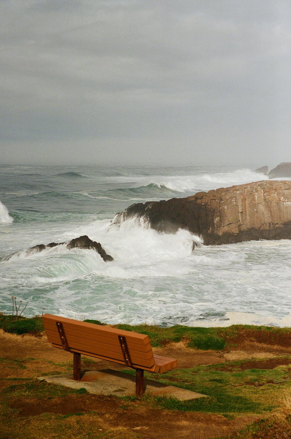 A wooden bench overlooks a rocky coastline with large waves crashing against the shore under a cloudy sky.