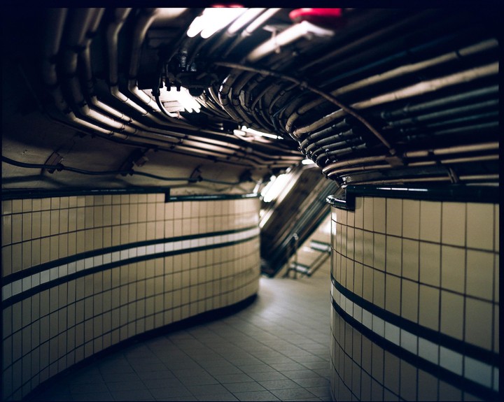 A curved subway tunnel features tiled walls and an exposed ceiling with numerous pipes and wires.