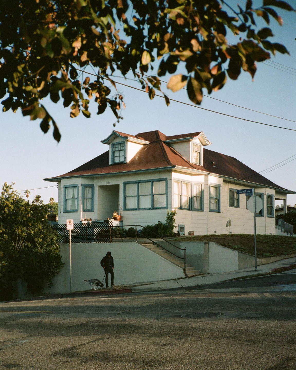 A man walks a dog on a leash in front of a white house with a red roof on a street corner.