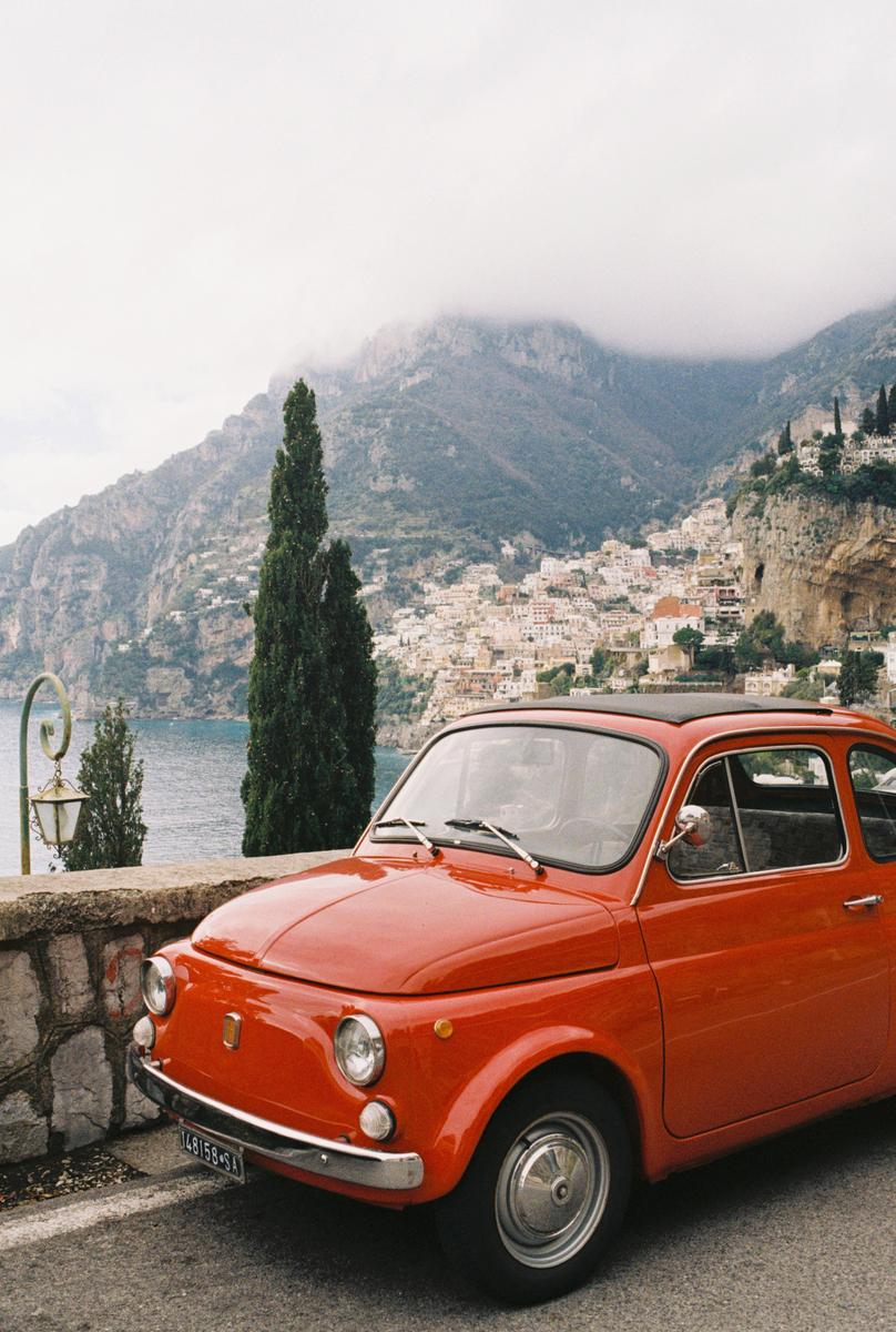 An orange car is parked on a road overlooking a coastal town and mountains.