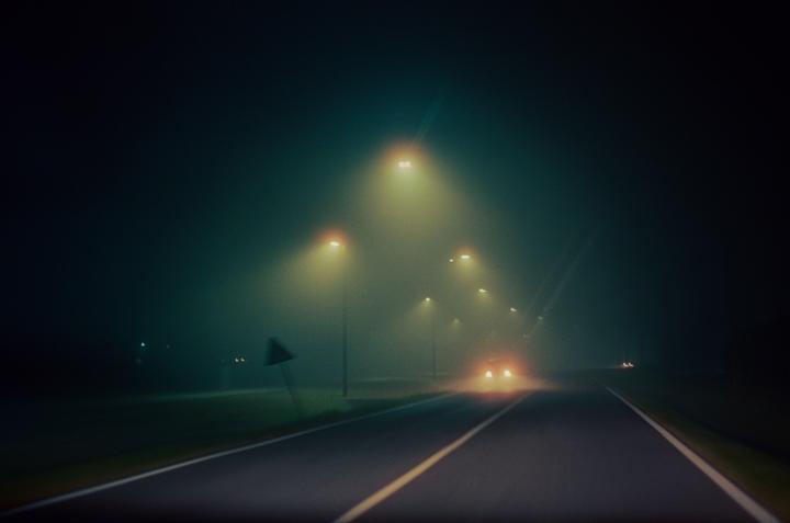 A car with its headlights on drives down a foggy road lined with streetlights at night.