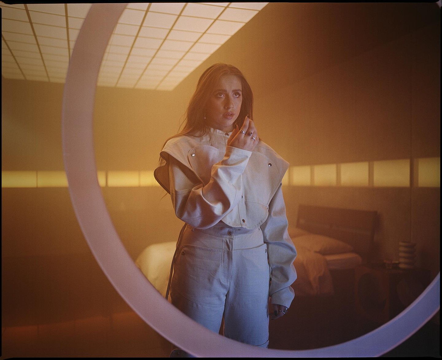 A woman stands in a room with a bed in the background, looking at her hand.