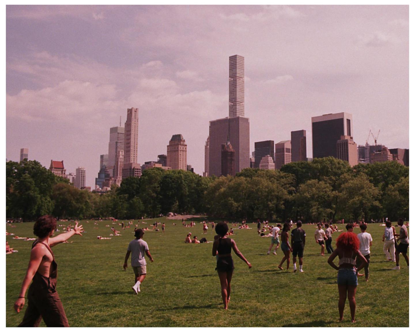 People are enjoying a summer day in Central Park with the New York City skyline in the background.