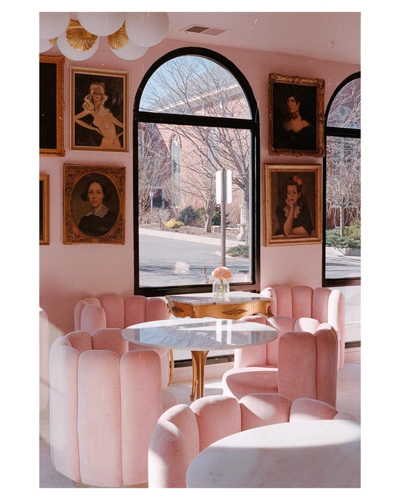 A cafe with pink chairs and tables is visible through an arched window, with paintings on the wall.