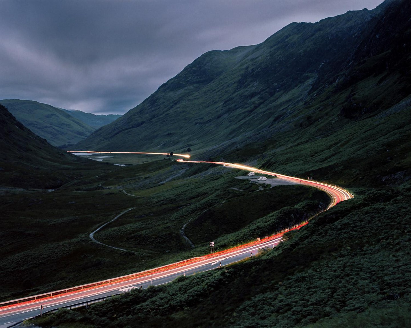 A long exposure of a winding road with light trails from vehicles through a mountain valley at dusk.