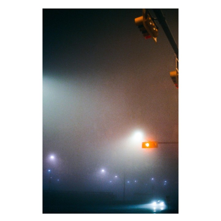 A street at night is obscured by thick fog, with a traffic light showing an orange signal and the headlights of a vehicle visible in the distance.
