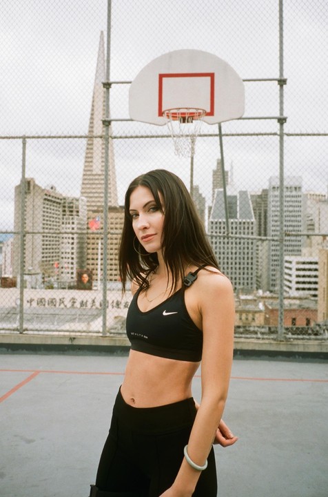 A woman stands on a rooftop basketball court with a city skyline in the background.