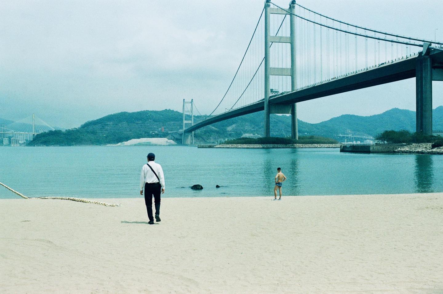 Two men stand on a sandy beach with a large suspension bridge and mountains in the background.