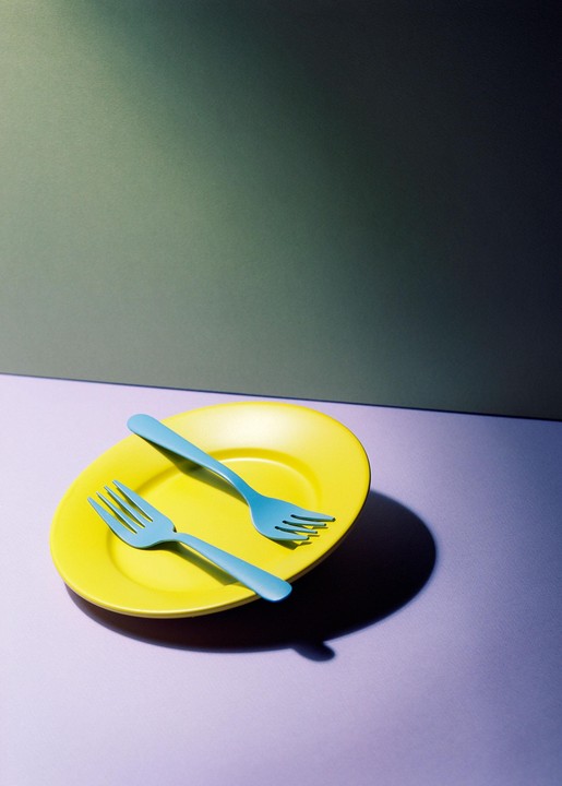 Two blue forks rest on a yellow plate, casting shadows on a purple surface with a green background.