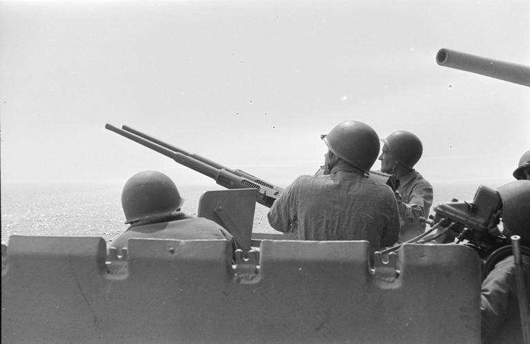 Several men in military helmets operate a large gun on a ship, looking out over the ocean.