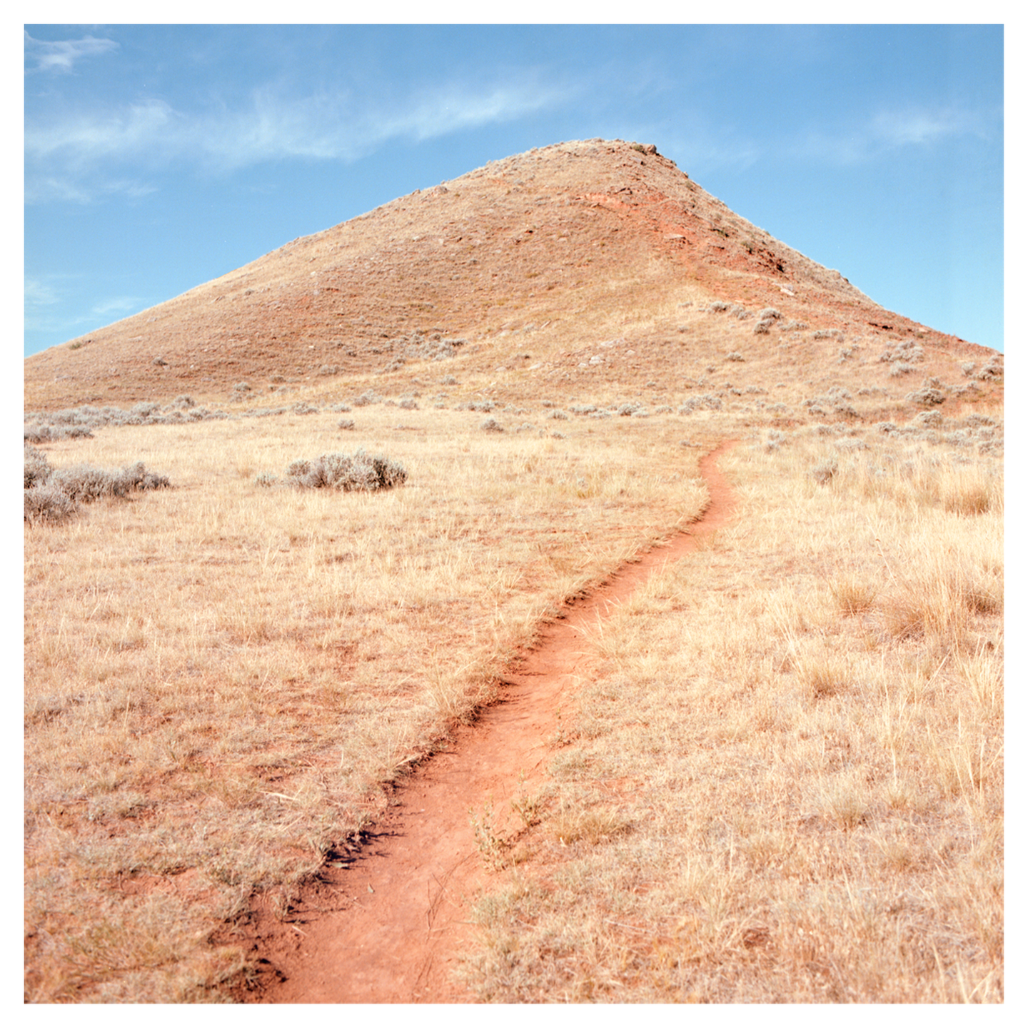 A narrow dirt path leads through dry grass towards a small hill under a blue sky.