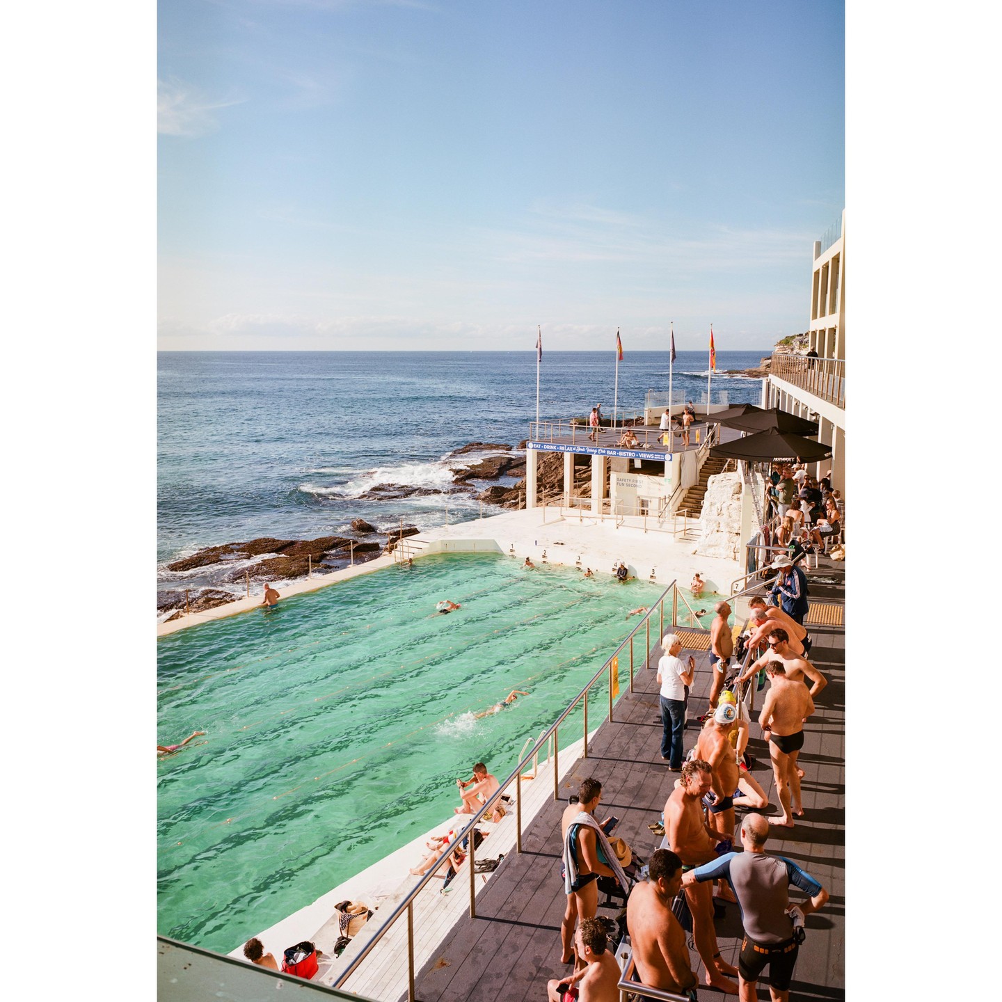 A swimming pool next to the ocean is filled with people swimming and sunbathing.