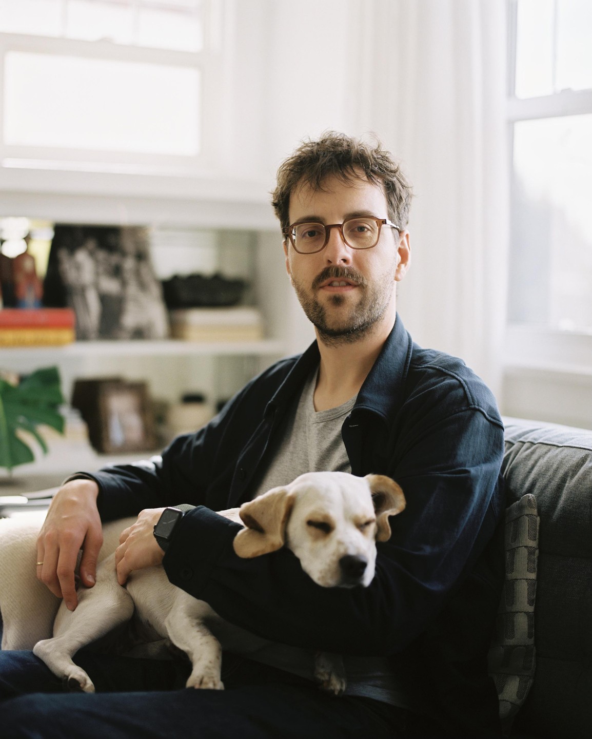 A man with glasses sits on a sofa, holding a small dog with closed eyes.