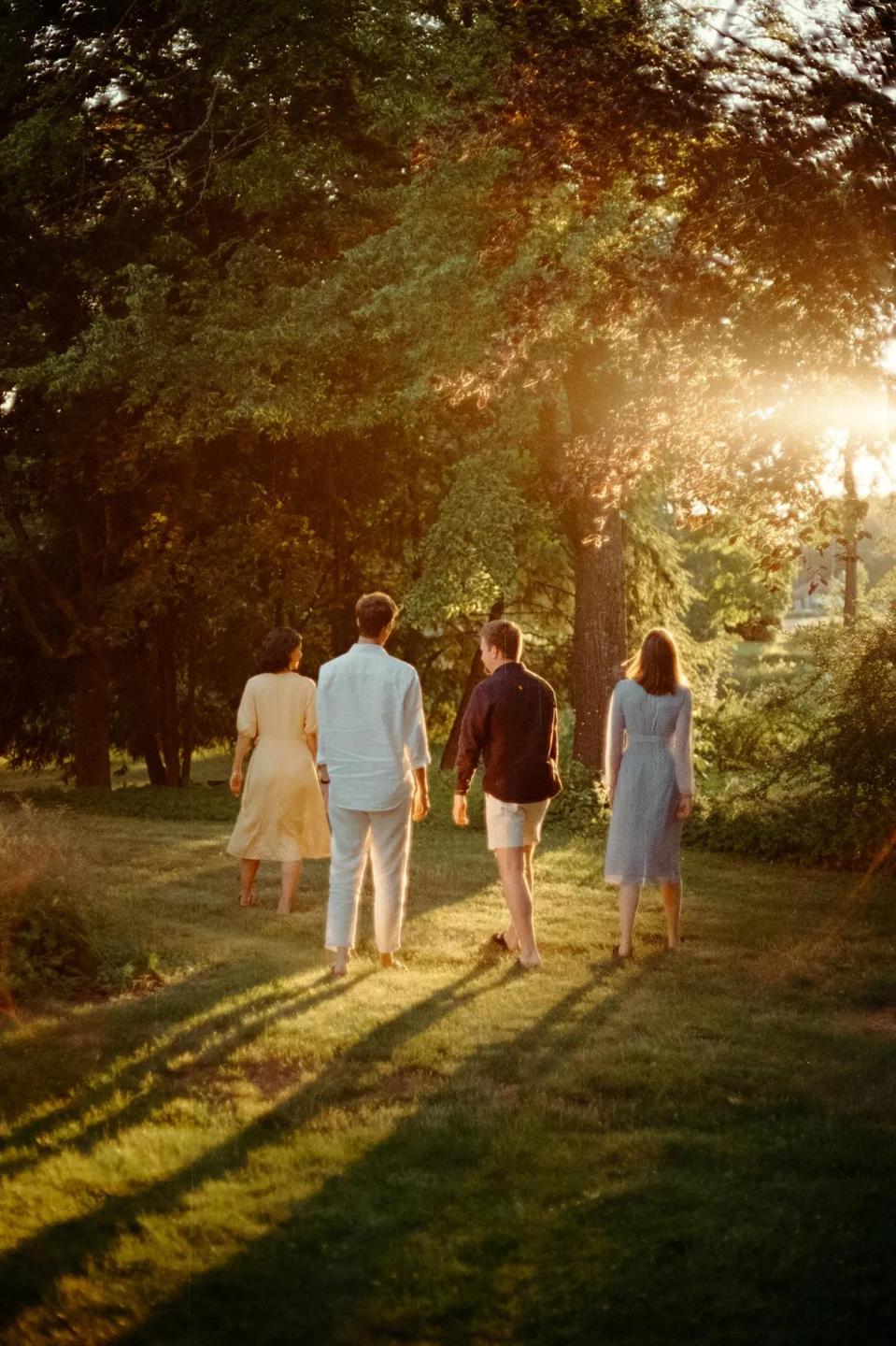 Four people walk barefoot through a grassy area with trees and golden hour sunlight.