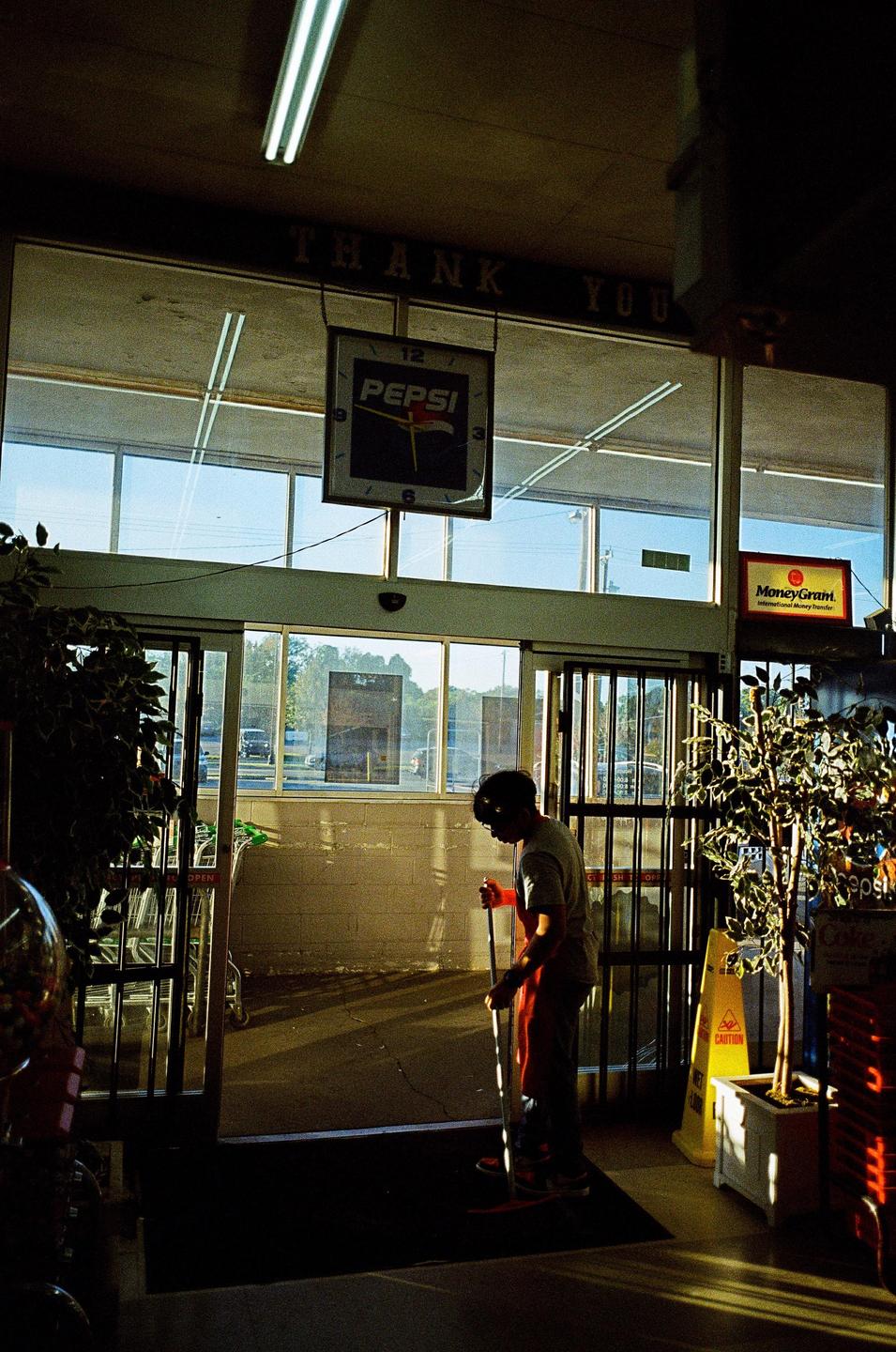 A man sweeps the floor inside a store entrance during golden hour, with a Pepsi clock and a MoneyGram sign visible through the glass doors.