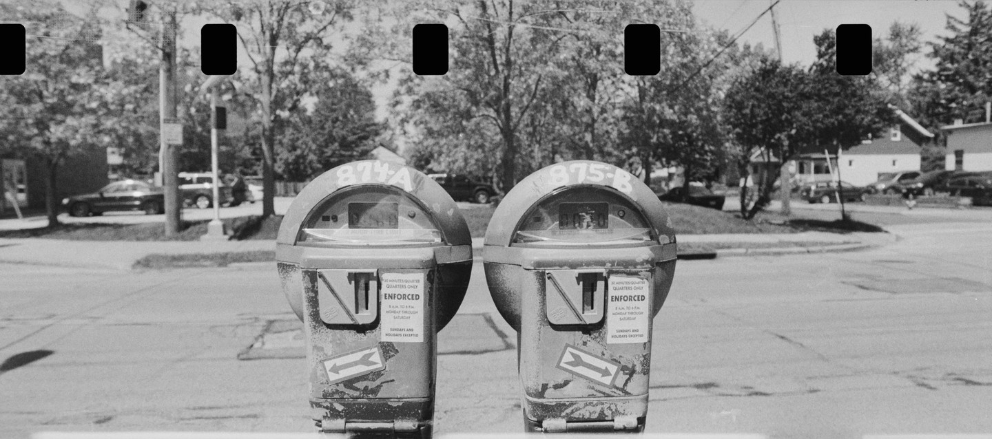 Two parking meters stand side by side on a street with trees and houses in the background.