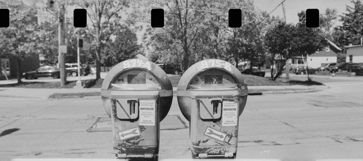 Two parking meters stand side by side on a street with trees and houses in the background.