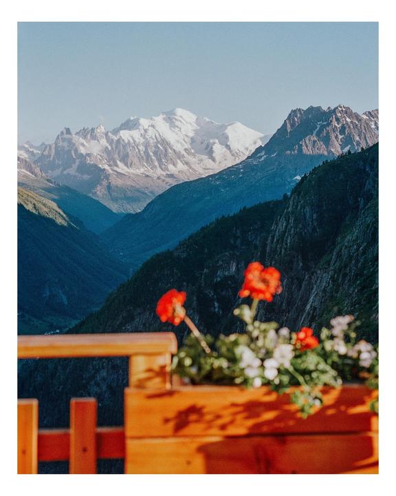 A mountain range with snow-capped peaks is visible behind a wooden balcony with red and white flowers in a planter.
