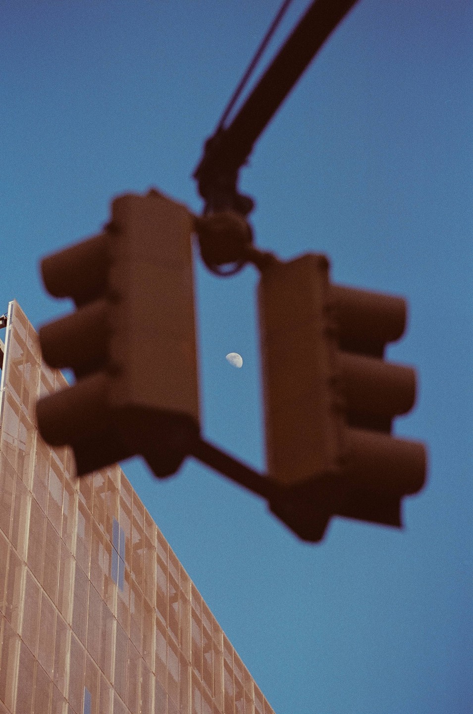A crescent moon is visible in the blue sky between two out-of-focus traffic lights, with a building on the left.