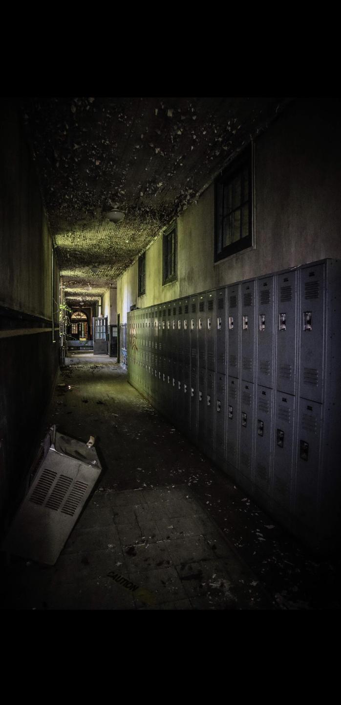 A long, dark hallway in an abandoned school is lined with lockers and illuminated by light from windows and the far end.