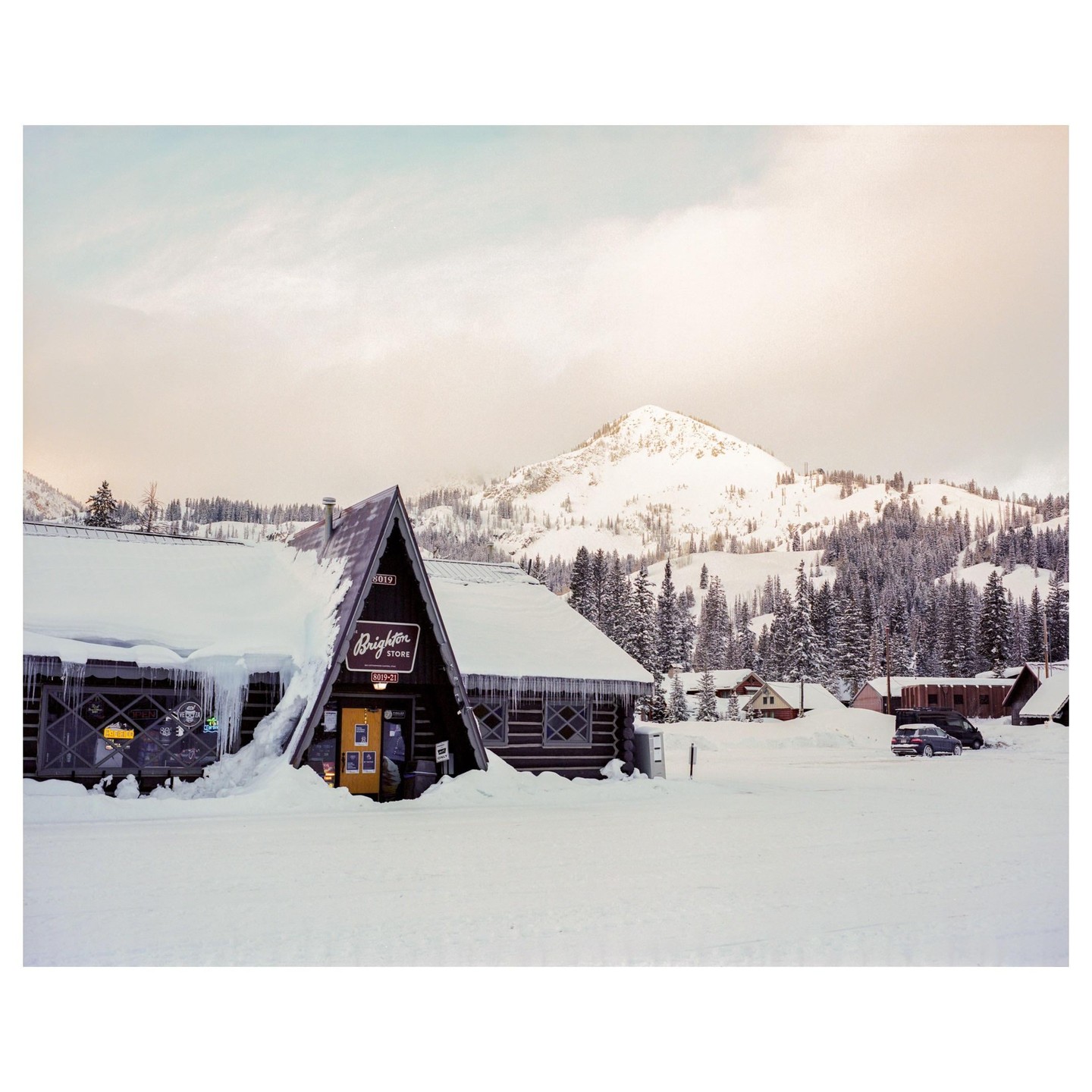 A snow-covered A-frame building with icicles hanging from its roof stands in a snowy landscape with mountains and pine trees in the background.