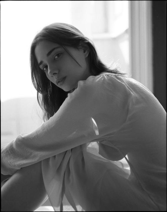A woman with long hair sits with her knees drawn up, looking over her shoulder at the viewer.