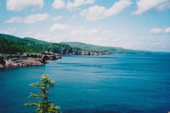 A body of water with a rocky shoreline and a tree in the foreground is visible under a blue sky with clouds.