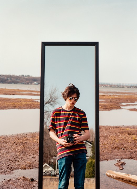 A young man wearing googly eye glasses holds a camera while standing in front of a mirror outdoors.