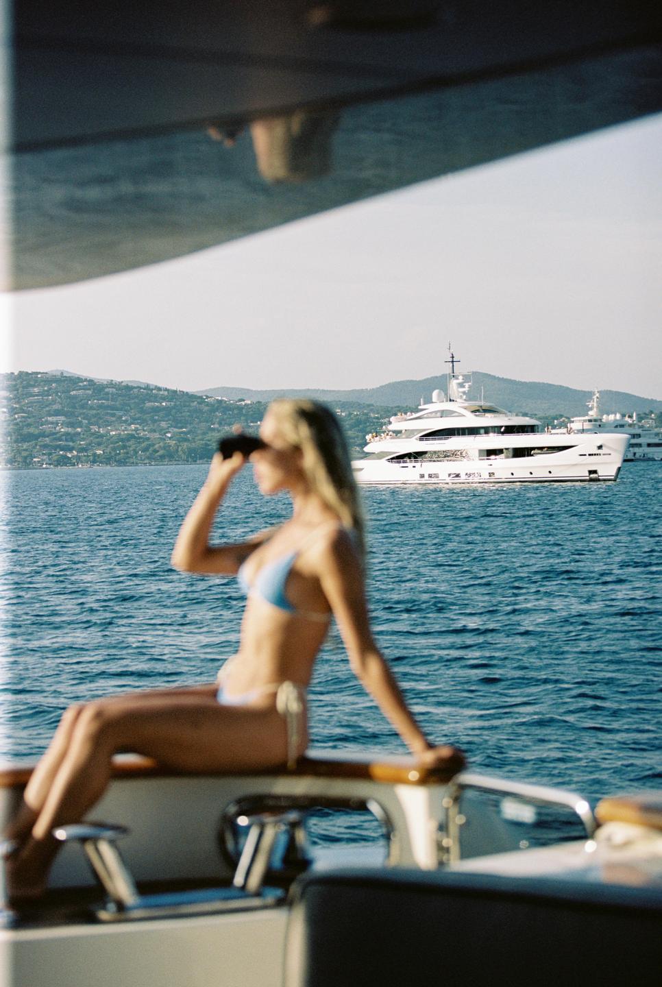 A woman in a bikini looks through binoculars from a boat at a yacht on the water.