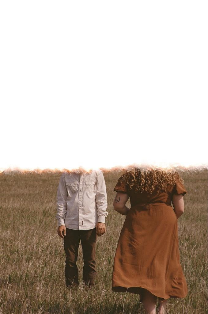 A man and a woman stand in a field of tall grass with their heads obscured by a bright white light.