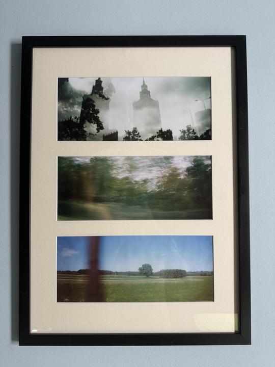 Three framed photographs are displayed vertically on a wall, with the top image showing a hazy city building, the middle image showing blurred green foliage, and the bottom image showing a green field with a single tree.