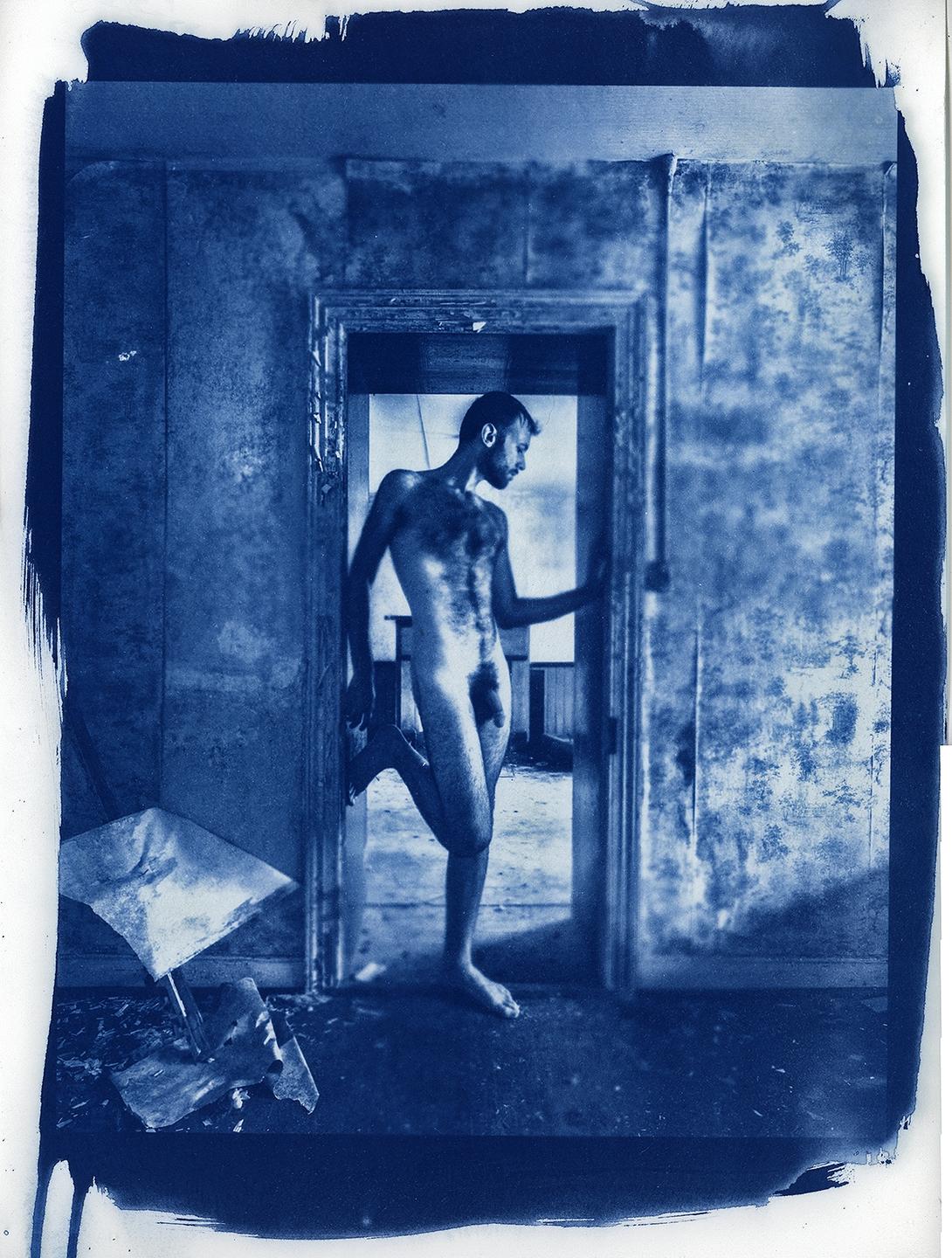 A nude man stands in a doorway in a derelict room, with a broken umbrella on the floor.